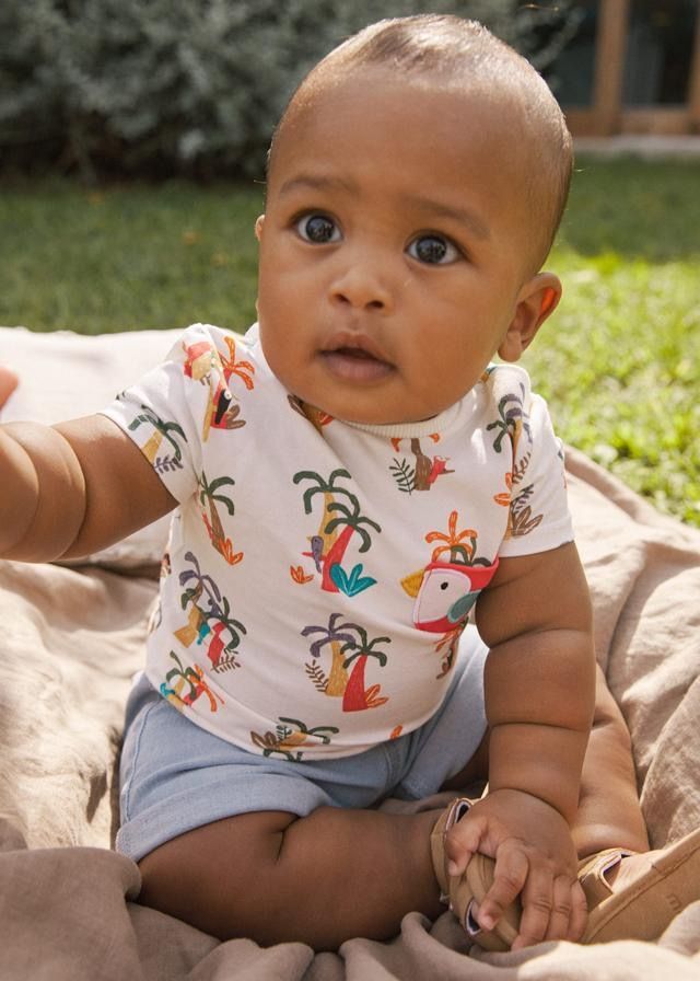 Un bambino seduto su una coperta all'aperto, con indosso una maglietta bianca con disegni colorati di palme e pantaloncini blu, guarda in alto.