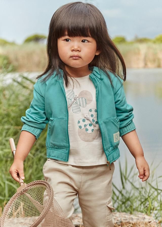 Bambino con giacca verde acqua, camicia beige e pantaloni color cachi vicino all'acqua, con in mano una retina.