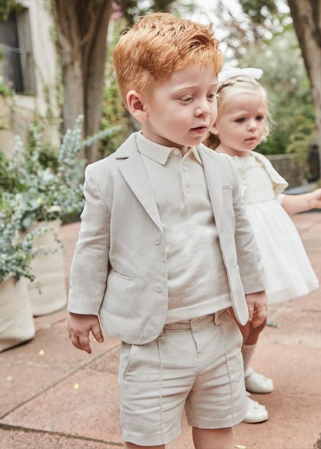 Ragazzo in abito marrone chiaro, che guarda di lato. Ragazza in abito bianco dietro di lui. Scenario all'aperto.