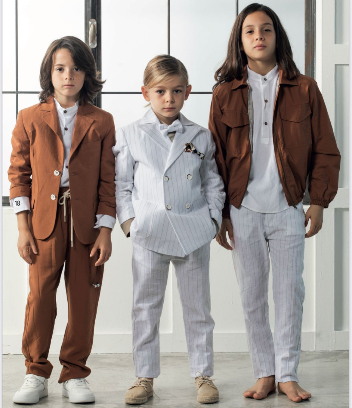Tre ragazzi in abiti formali posano davanti a una finestra. Uno indossa un abito marrone, uno un abito gessato bianco e uno una giacca marrone.