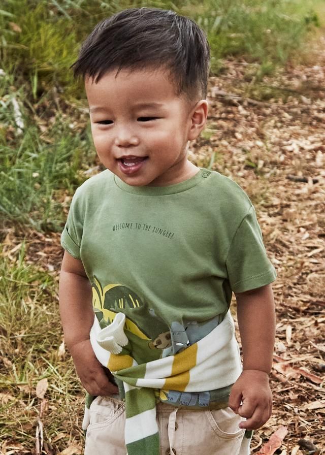 Bambino sorridente, con indosso una maglietta verde e pantaloni beige, in un ambiente esterno.