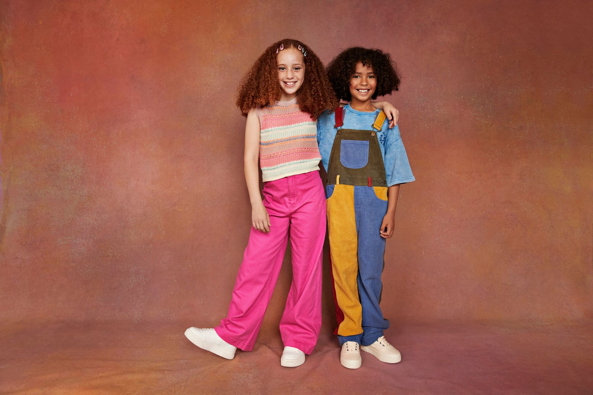 Due bambini in abiti colorati, in posa. Uno con pantaloni rosa e l'altro con una tuta rattoppata, entrambi sorridenti.