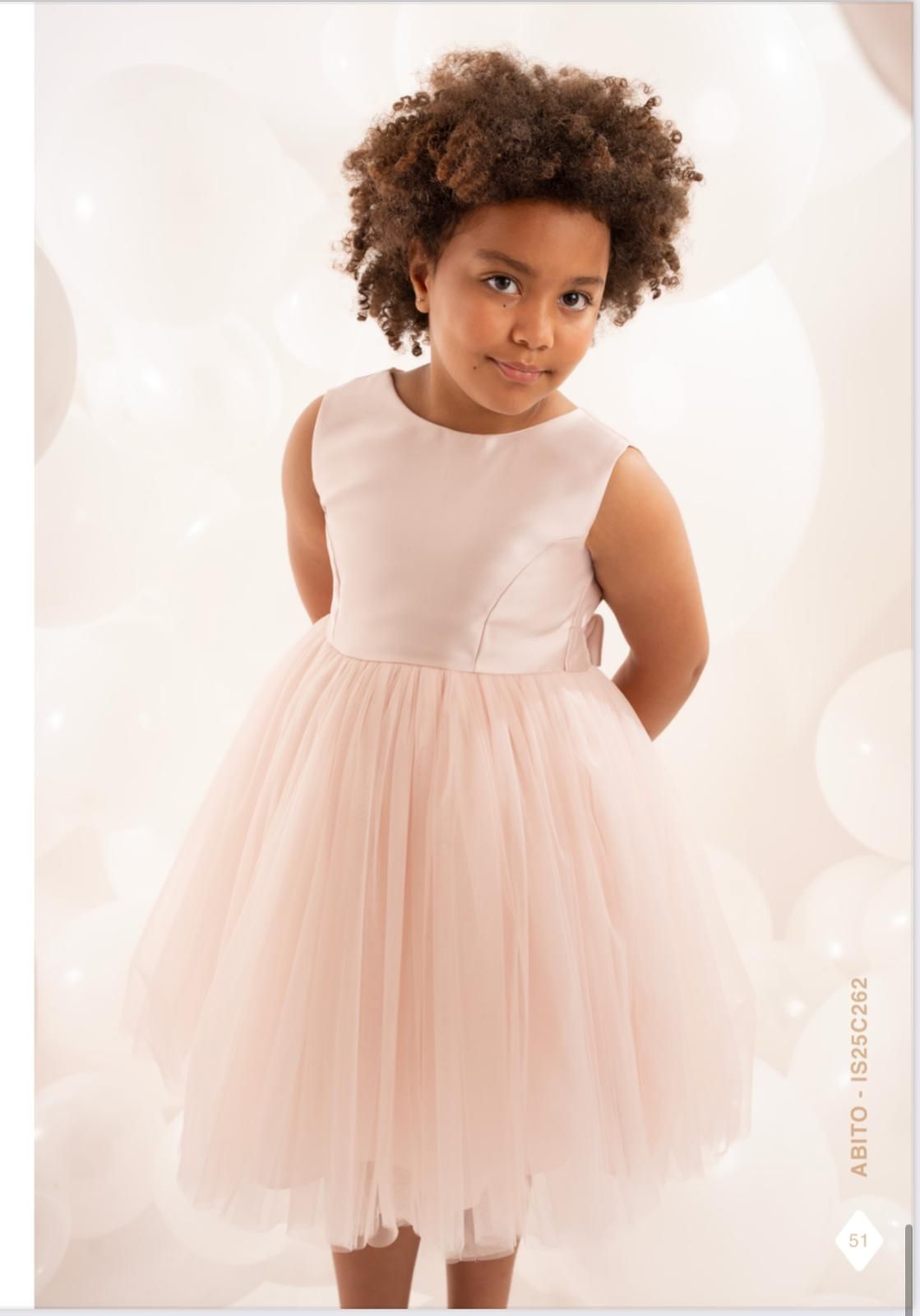 Una ragazzina con un vestito rosa, le mani dietro la schiena, davanti a palloncini bianchi.