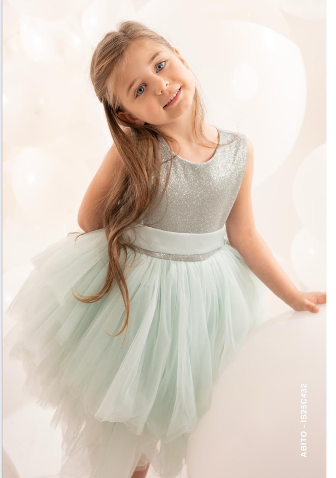 Ragazza con un vestito scintillante color argento e verde menta, sorridente alla telecamera, appoggiata a una superficie bianca, con palloncini sullo sfondo.