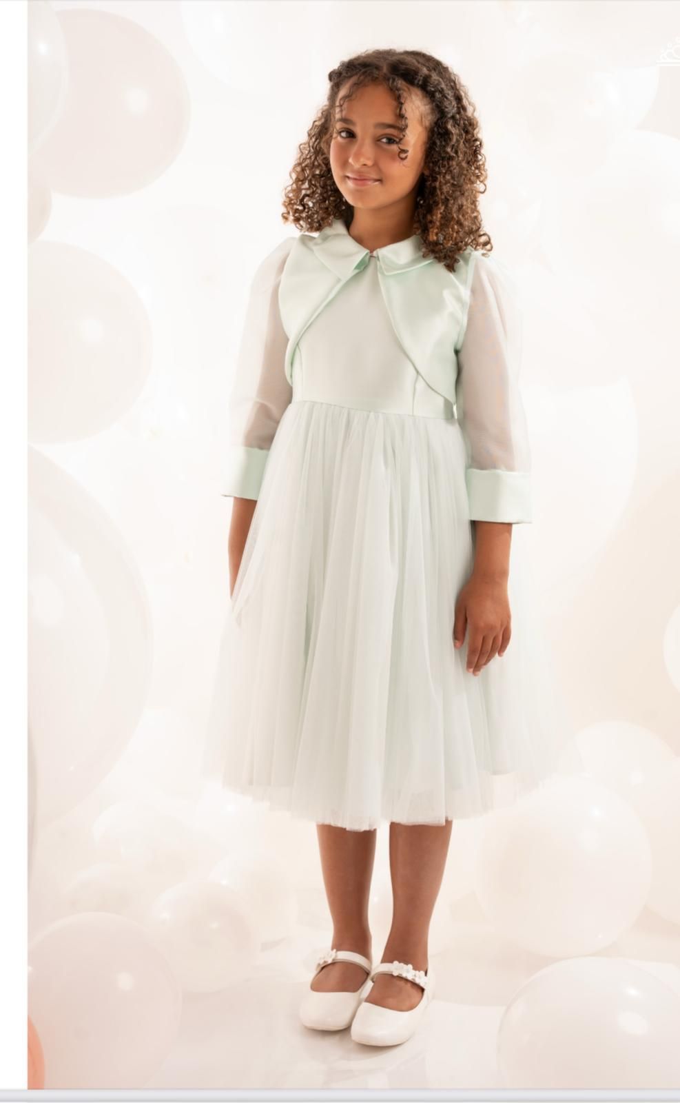 Una ragazzina indossa un vestito verde chiaro e una giacca coordinata, in piedi tra i palloncini.