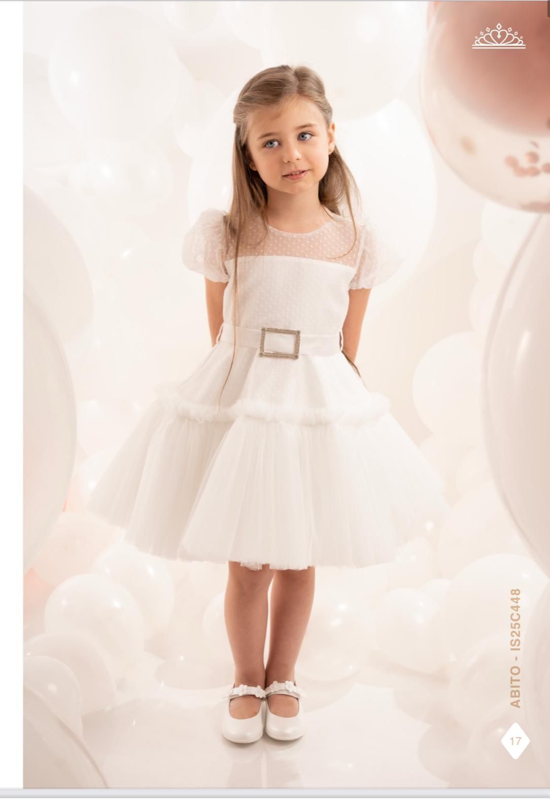 Ragazza in abito bianco con maniche a sbuffo, in piedi, che guarda la telecamera. Sfondo con palloncini bianchi.