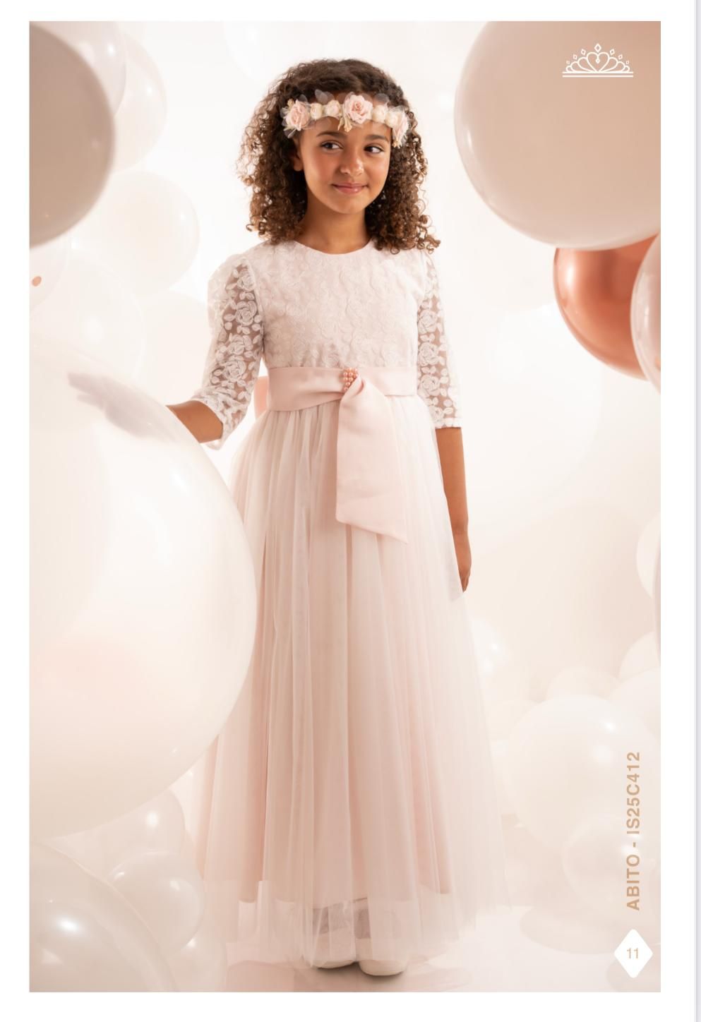 Ragazza con abito bianco, maniche in pizzo e corona di fiori, circondata da palloncini.