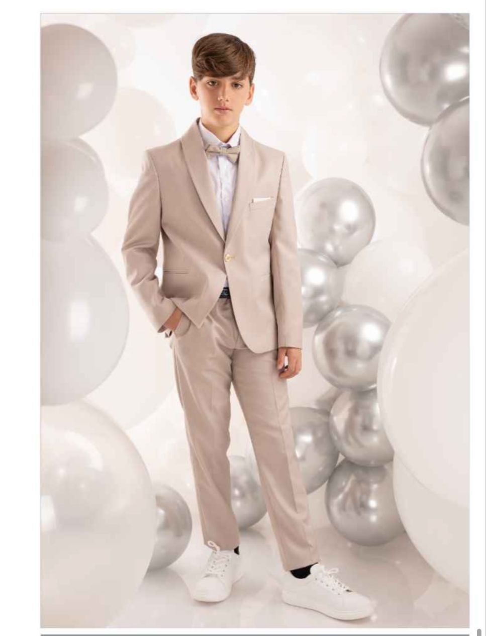 Un ragazzo in abito marrone chiaro, papillon, scarpe da ginnastica, circondato da palloncini bianchi e argento.
