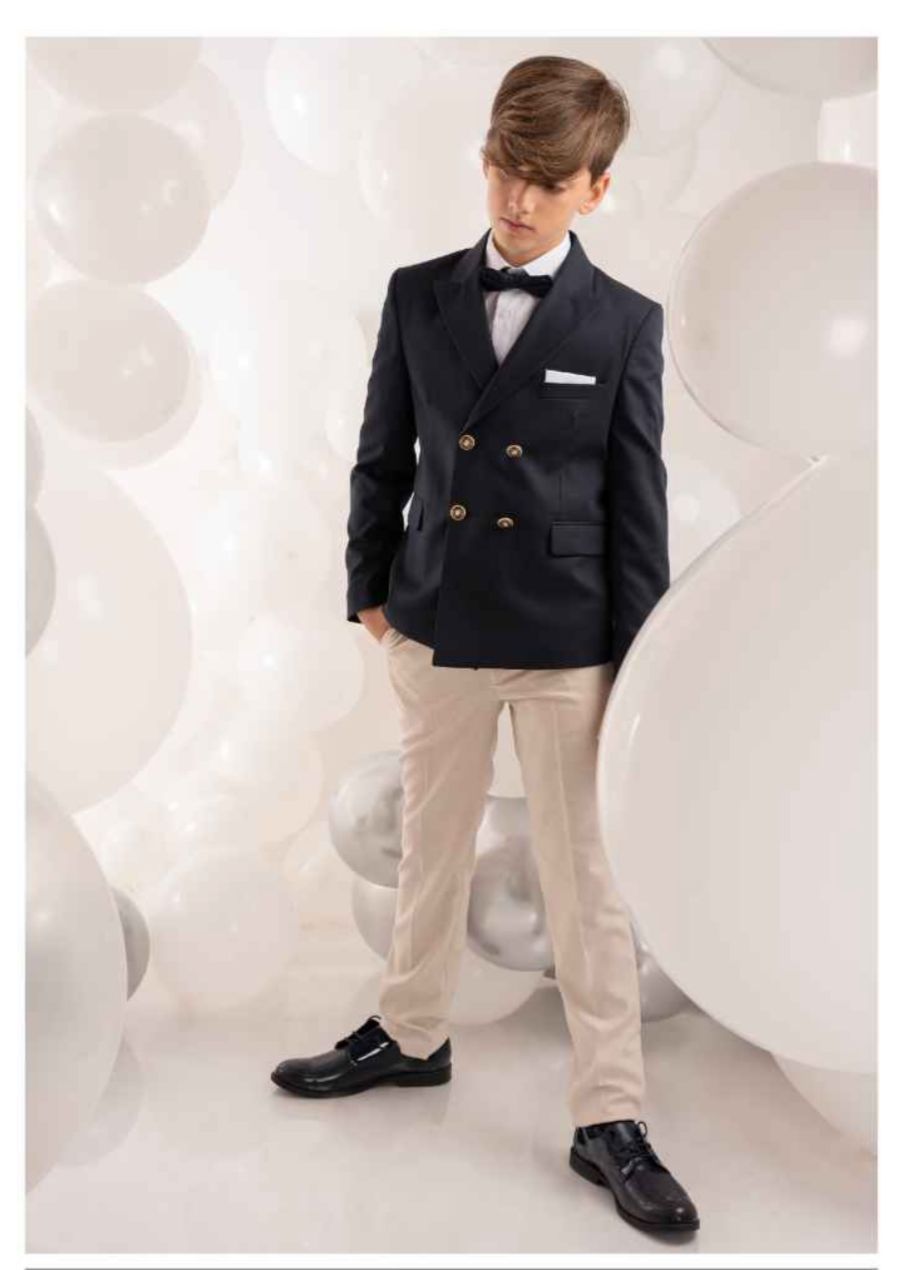 Un ragazzo con una giacca blu navy, pantaloni beige e un papillon, in piedi davanti a palloncini bianchi.