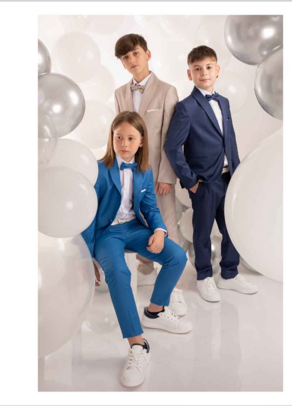 Tre bambini in giacca e cravatta posano con dei palloncini; giacca e cravatta blu, beige e blu navy. Sfondo bianco.