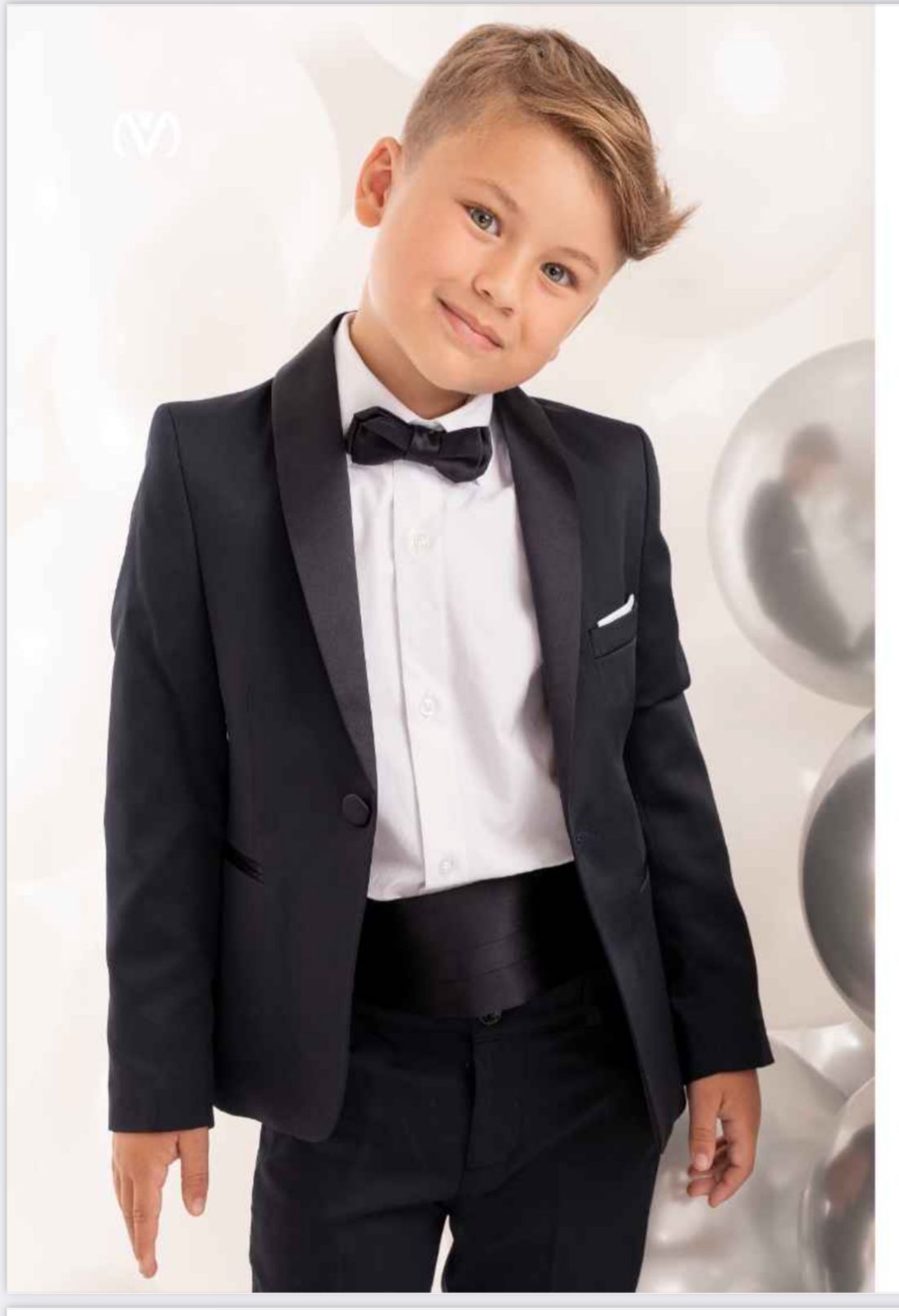 Un ragazzo in smoking nero con papillon, sorridente, in piedi davanti a dei palloncini.