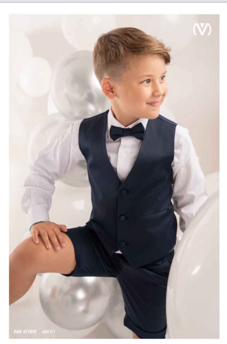 Un ragazzo con un gilet e pantaloncini blu, una camicia bianca e un papillon, circondato da palloncini argentati.