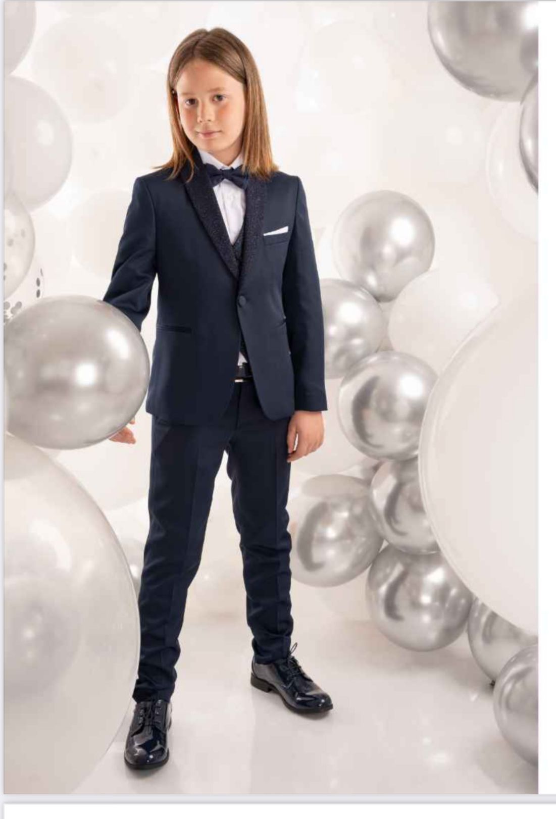 Ragazzo in abito blu navy con papillon, che tiene in mano un palloncino argentato, circondato da palloncini argentati e bianchi.