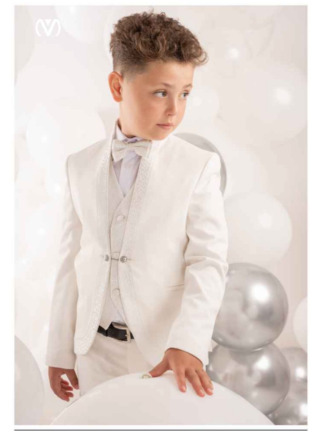 Ragazzo in abito bianco e papillon, in piedi davanti a palloncini bianchi e argentati.