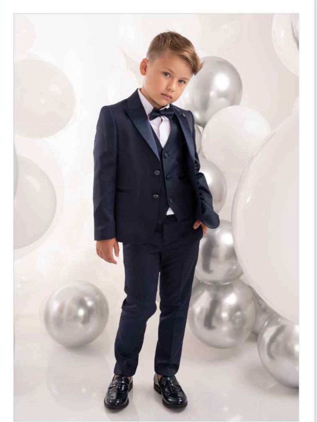 Un ragazzo in abito blu scuro con papillon, in piedi davanti a palloncini bianchi e argentati.
