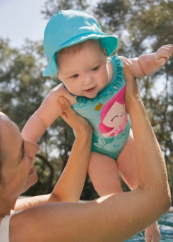 Un bambino con un costume da bagno verde acqua e un cappello da sole blu è tenuto in braccio da un adulto; sorrisi, ambiente soleggiato all'aperto.
