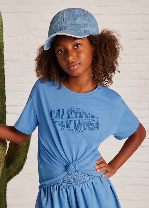 Una ragazza con una maglietta blu e un berretto di jeans posa vicino a un cactus. Sulla maglietta c'è scritto