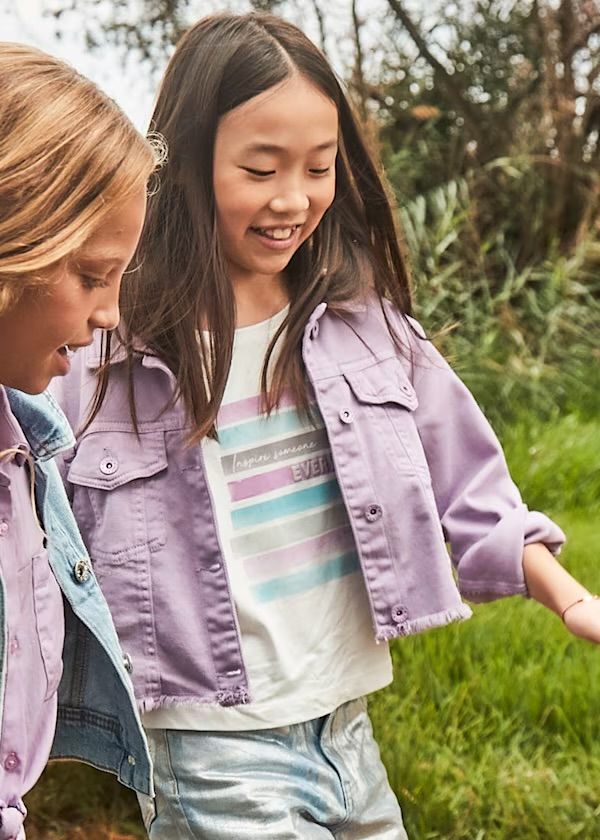 Due ragazze all'aperto; una indossa una giacca e una maglietta color lavanda, pantaloni argentati; sorridono.
