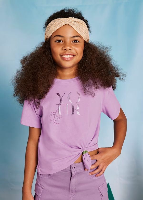 Ragazza con abito viola, fascia per capelli, maglietta annodata e pantaloncini, sorride.