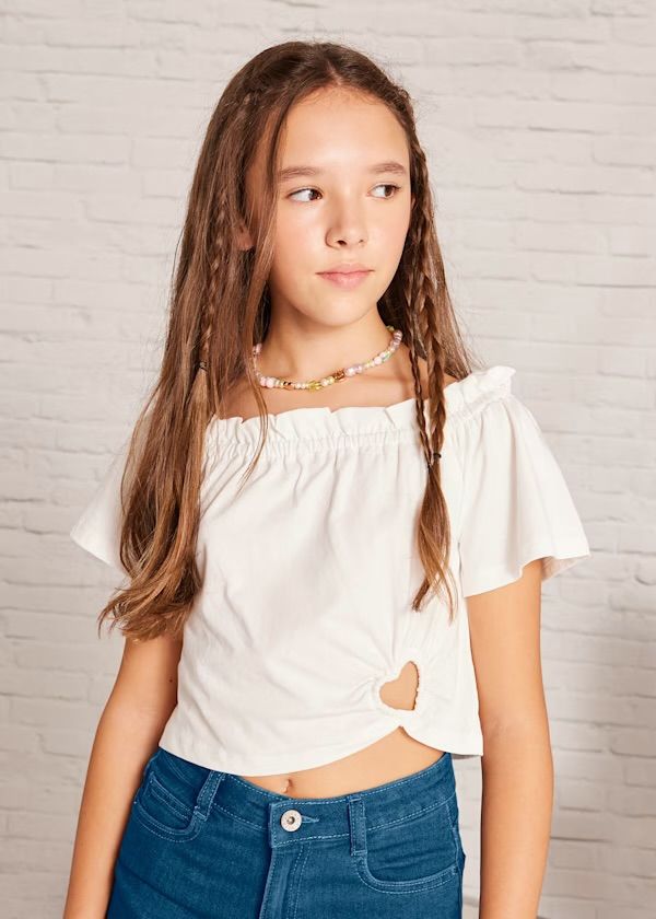 Ragazza che indossa un top bianco con spalle scoperte e un cuore ritagliato e jeans blu, in posa davanti a un muro di mattoni bianchi.