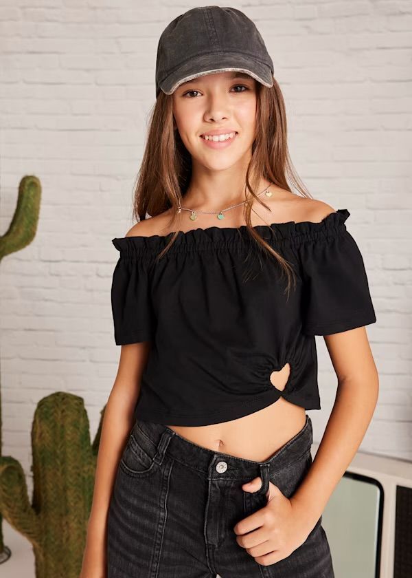 Ragazza che indossa un top nero con le spalle scoperte, un berretto e pantaloncini di jeans. Posa davanti a un muro di mattoni bianchi.