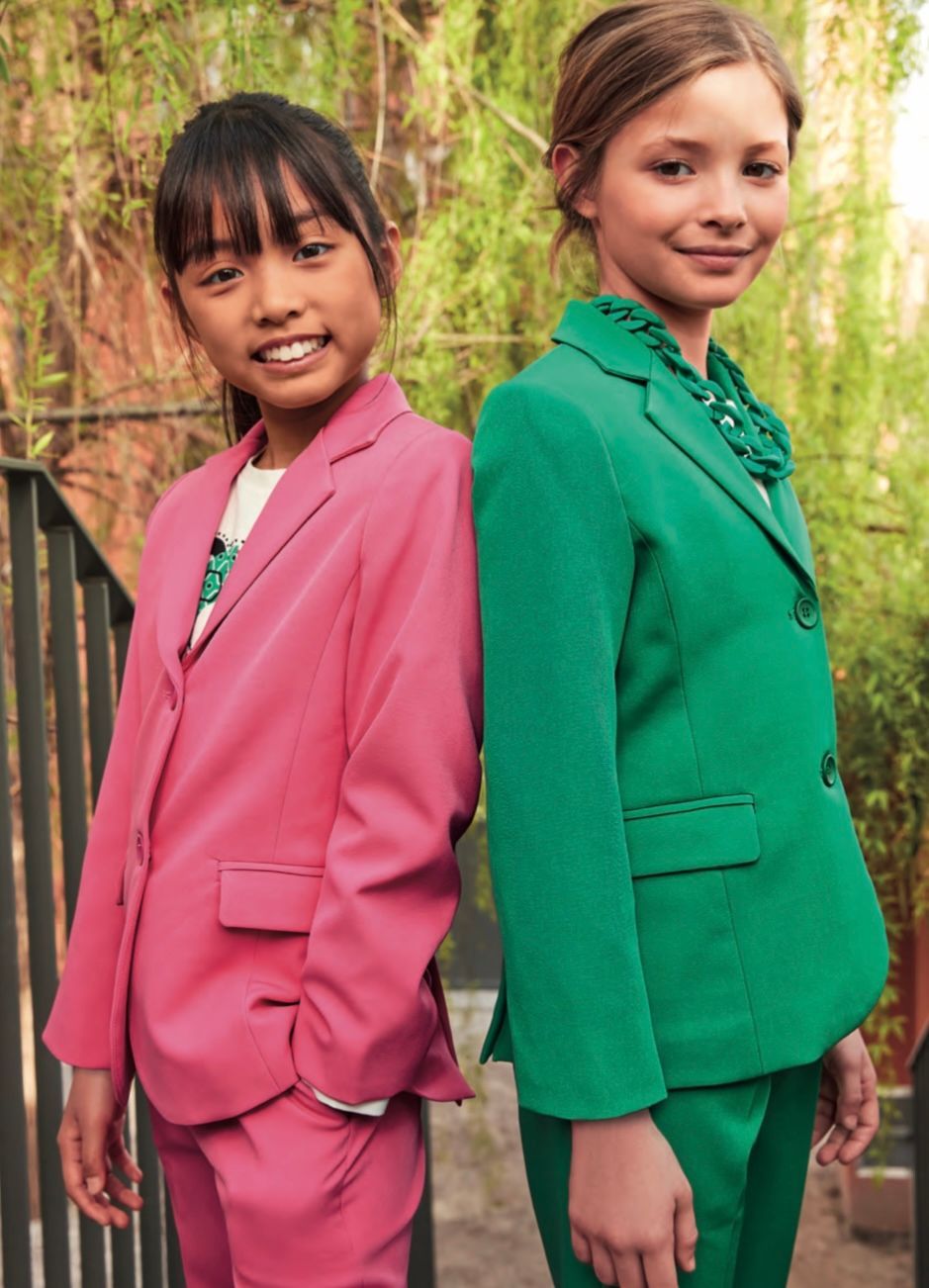 Due ragazze in abiti colorati sono in piedi, schiena contro schiena, fuori. Una indossa un abito rosa, l'altra un abito verde, entrambe sorridenti.