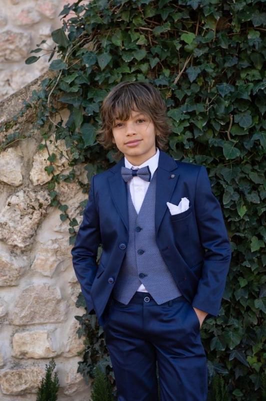 Ragazzo in abito blu navy con papillon e fazzoletto da taschino, in posa davanti a un muro di mattoni ricoperto di edera.