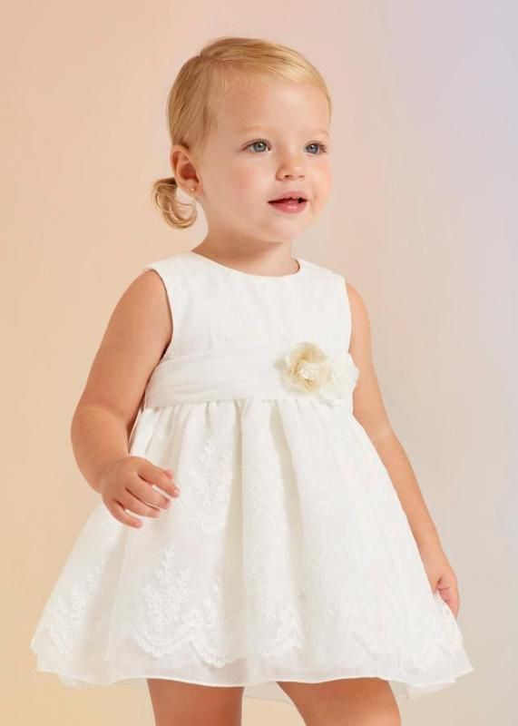 Una bambina bionda con un vestito bianco decorato con un fiore, posa davanti a uno sfondo sfumato e delicato.
