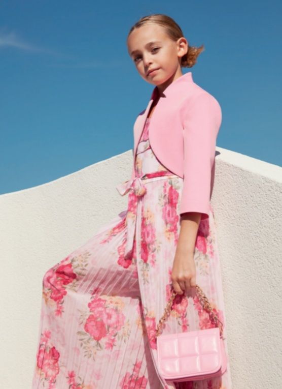 Ragazza con tuta e giacca a fiori rosa, che tiene in mano una borsa rosa, in posa contro un muro bianco e un cielo azzurro.