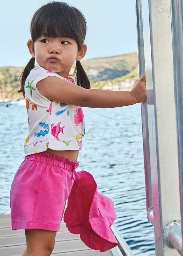 Bambino con pantaloncini rosa e maglietta a fiori, che tiene in mano un cappello, vicino all'acqua.