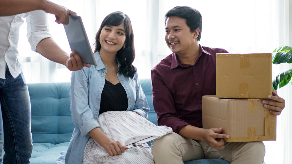 Couple smiles, holding boxes. Someone shows them a tablet. Indoors.