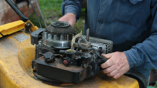 Man working on a yellow lawnmower engine. He is wearing a blue shirt, and is in an outdoor setting.