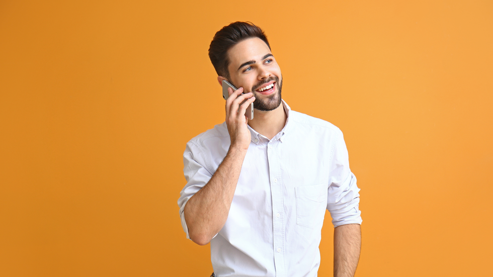 Man with a beard smiles while talking on a phone, wearing a white shirt against an orange background.