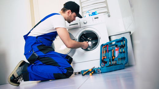 A repairman in blue overalls kneels by a washing machine, working with tools in an open case.
