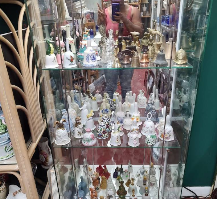 Glass display case with various bells on shelves, reflected person taking photo.