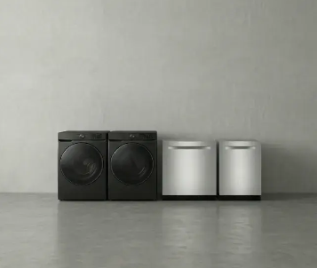 Two black front-loading laundry units stand next to two silver dishwashers against a light grey wall and concrete floor.