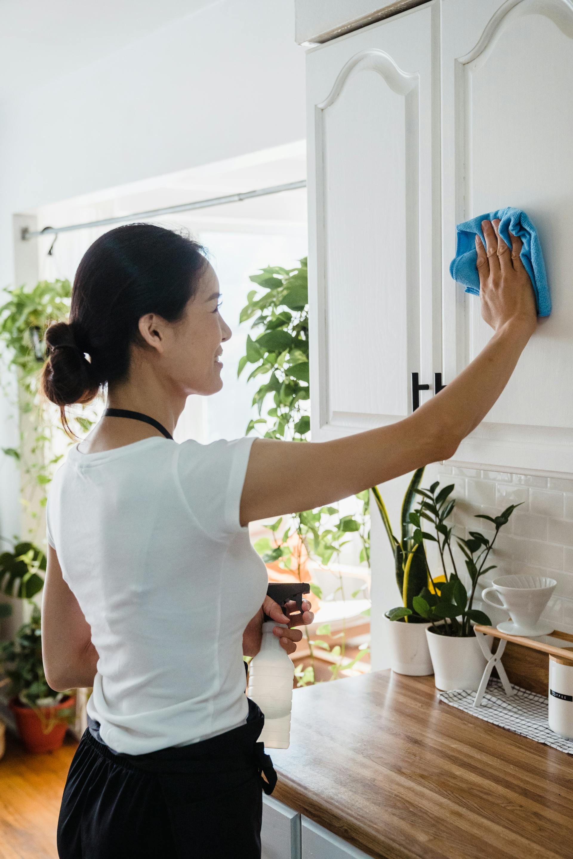 A woman is cleaning a kitchen cabinet with a blue cloth.