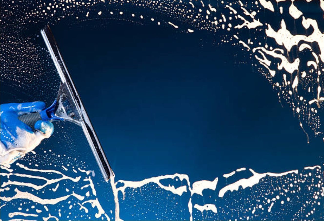 A blue-gloved hand uses a squeegee to wipe white soap suds off a dark blue glass surface.