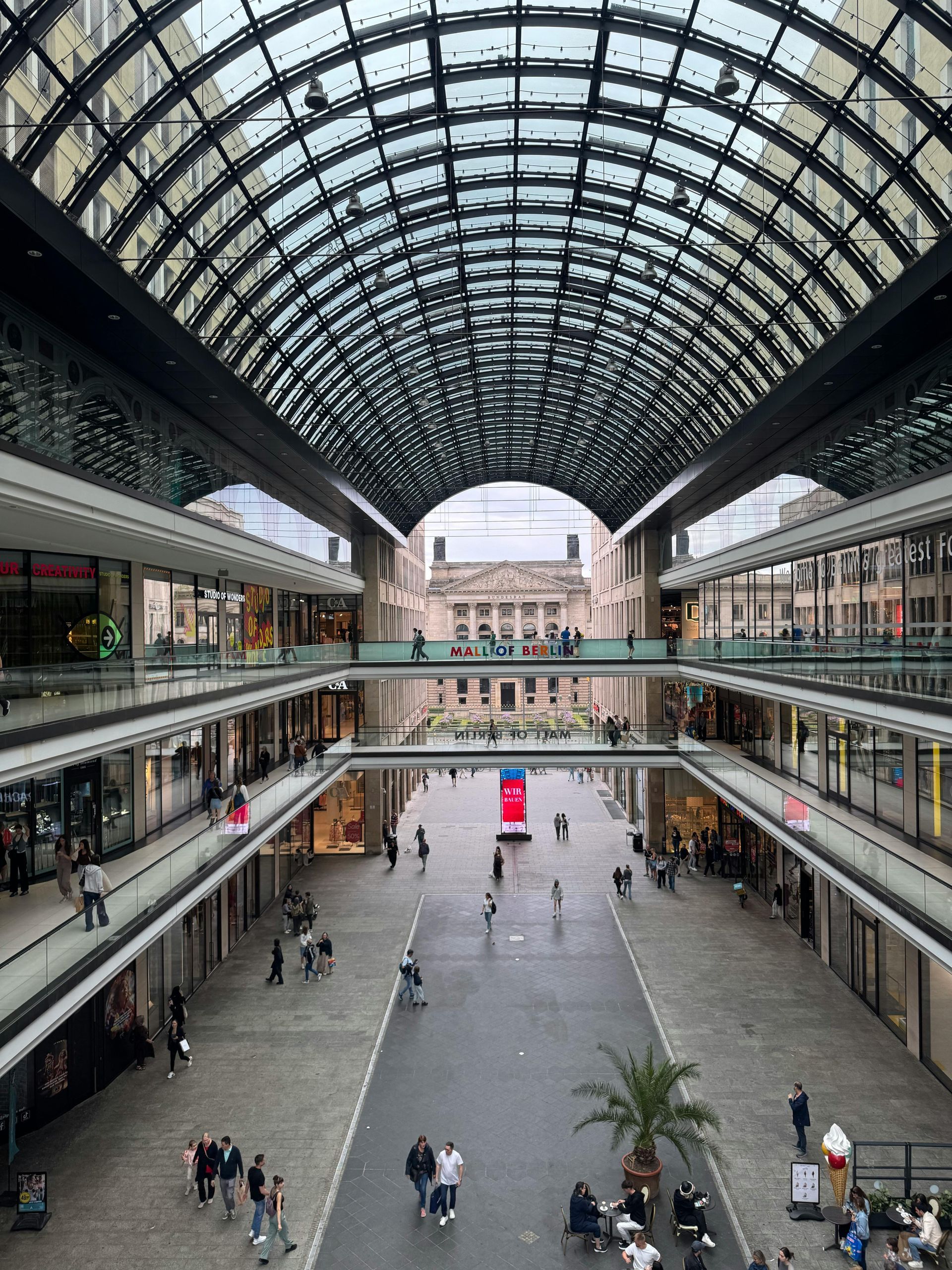 A multi-level shopping arcade with a high, vaulted glass ceiling and people walking on a stone-paved central ground floor.