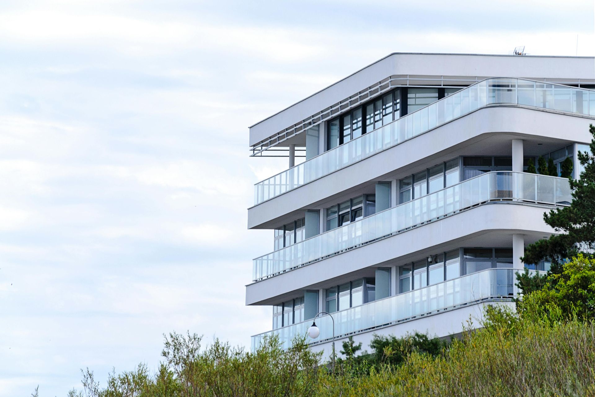 Modern white apartment building with glass balconies beside green trees under a cloudy sky