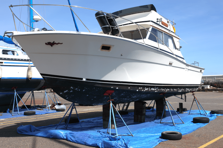 White motorboat on stands in a dry dock, blue bottom paint, against a blue tarp, on a sunny day.