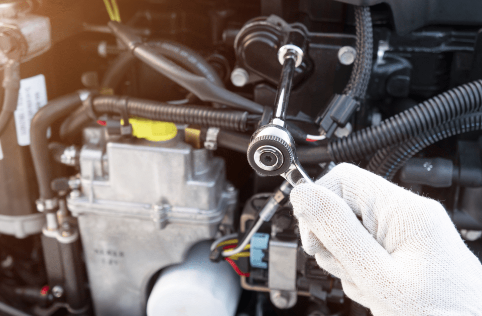 Gloved hand using a wrench on an engine; black hoses and wires surround metal components.