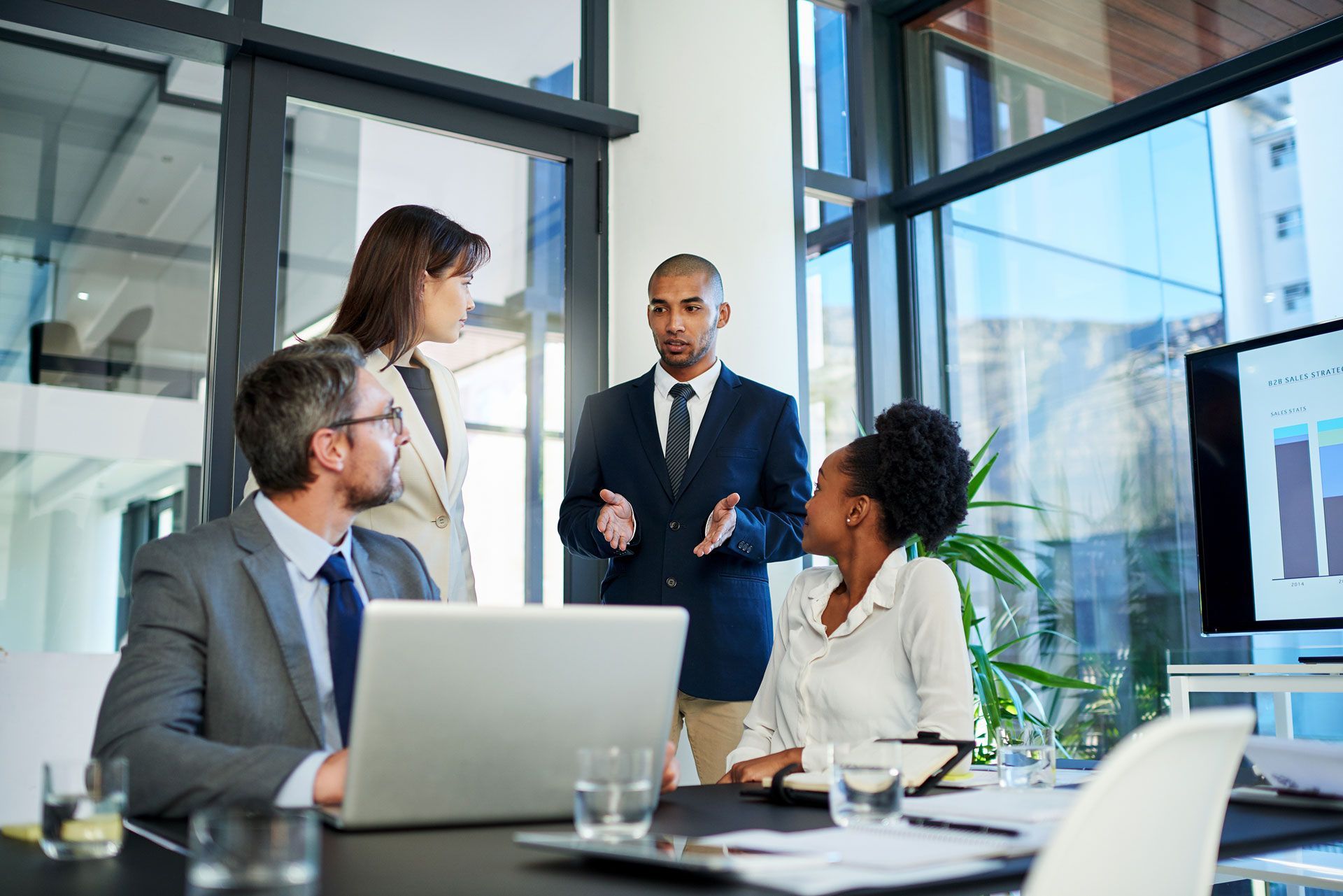 Business team in a modern office, gathered around a table; man speaking, laptop open, graph on screen.