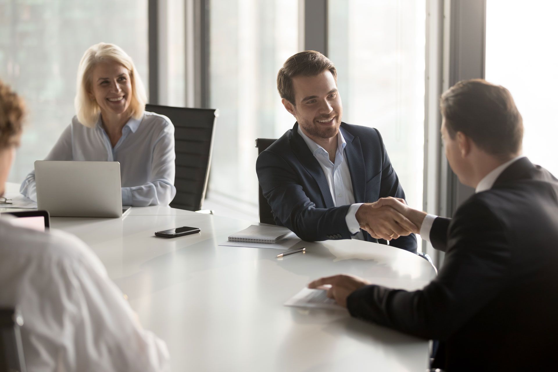 Business meeting with a handshake. Three people at a table; one is shaking hands. Window behind.