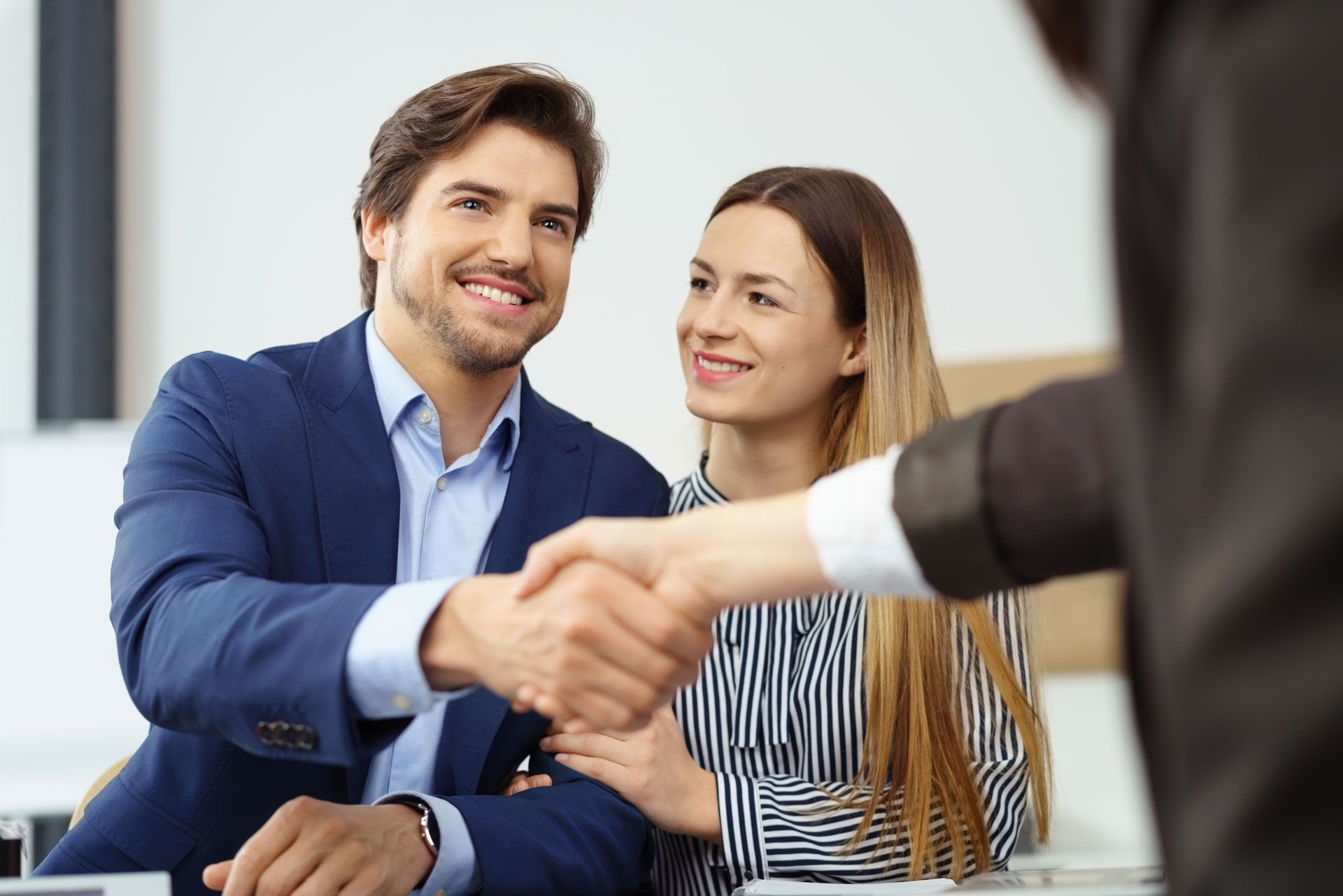 Man in blue suit shakes hands with person; woman smiles; office setting.