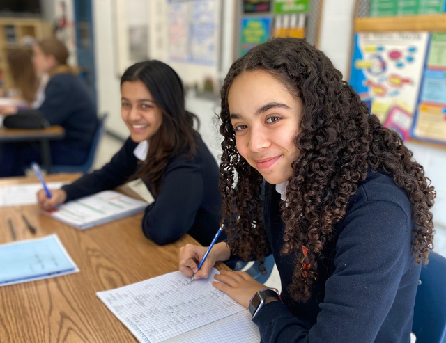 Two girls are sitting at a desk in a classroom writing in notebooks.