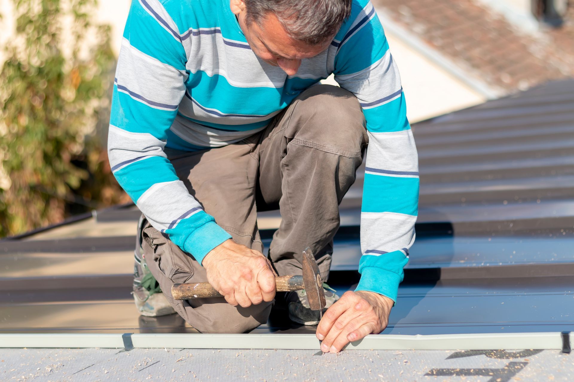 Roofer in orange hard hat measures roof tiles with a measuring tape; outdoors, sunny.