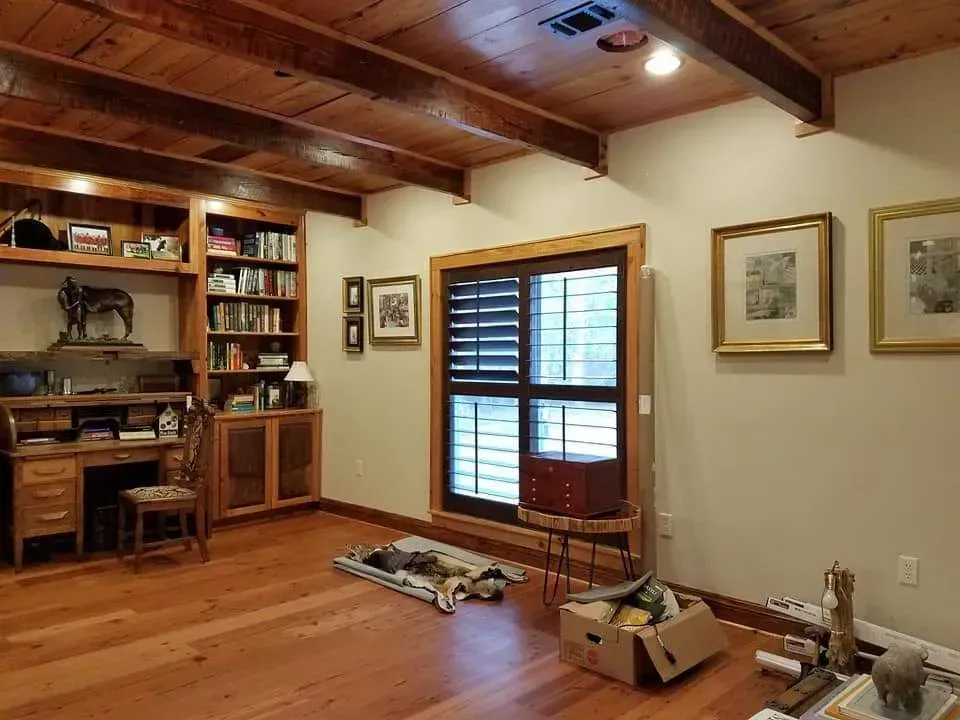 A living room with hardwood floors , a desk , shelves , and shutters.