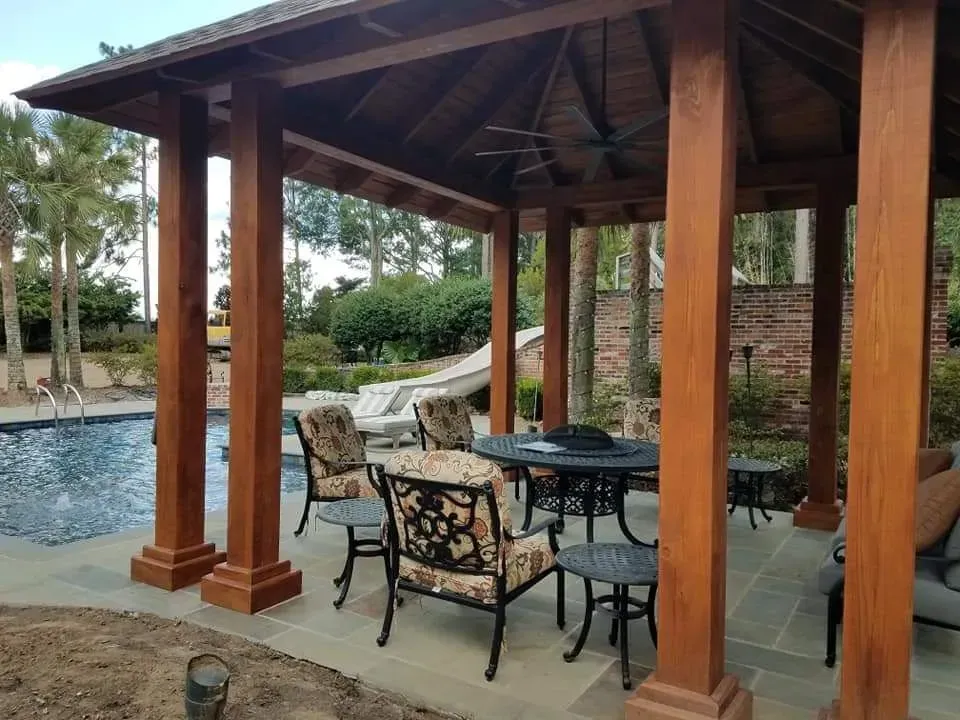 A wooden gazebo with a table and chairs in front of a pool.