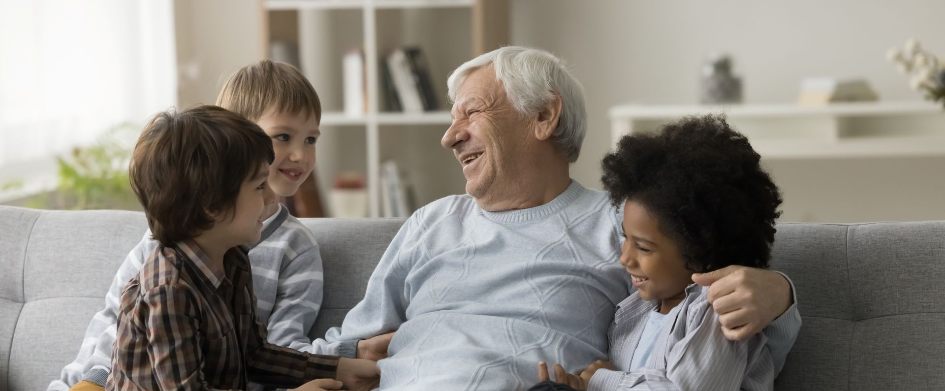 An older person sits with three children on a couch, all laughing. Interior setting.